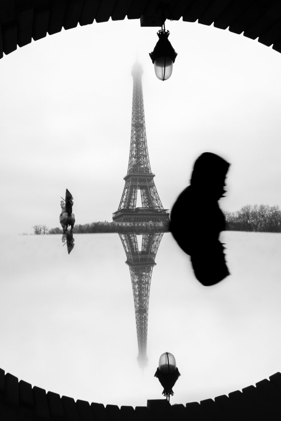 Silhouette of a man walking past in front of the Eiffel Tower, reflected in the water, monochromatic colouring.