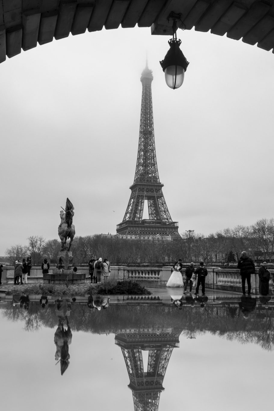 Eiffel Tower in the fog, reflected in a puddle, surrounded by people and buildings.