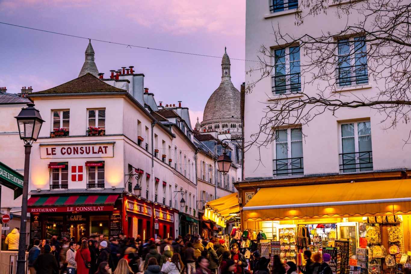 People on a lively street at sunset, with the Sacré-Cœur in the background.