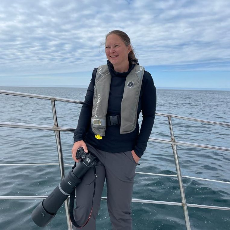 Woman with camera is standing on a boat, background with water and cloudy sky.
