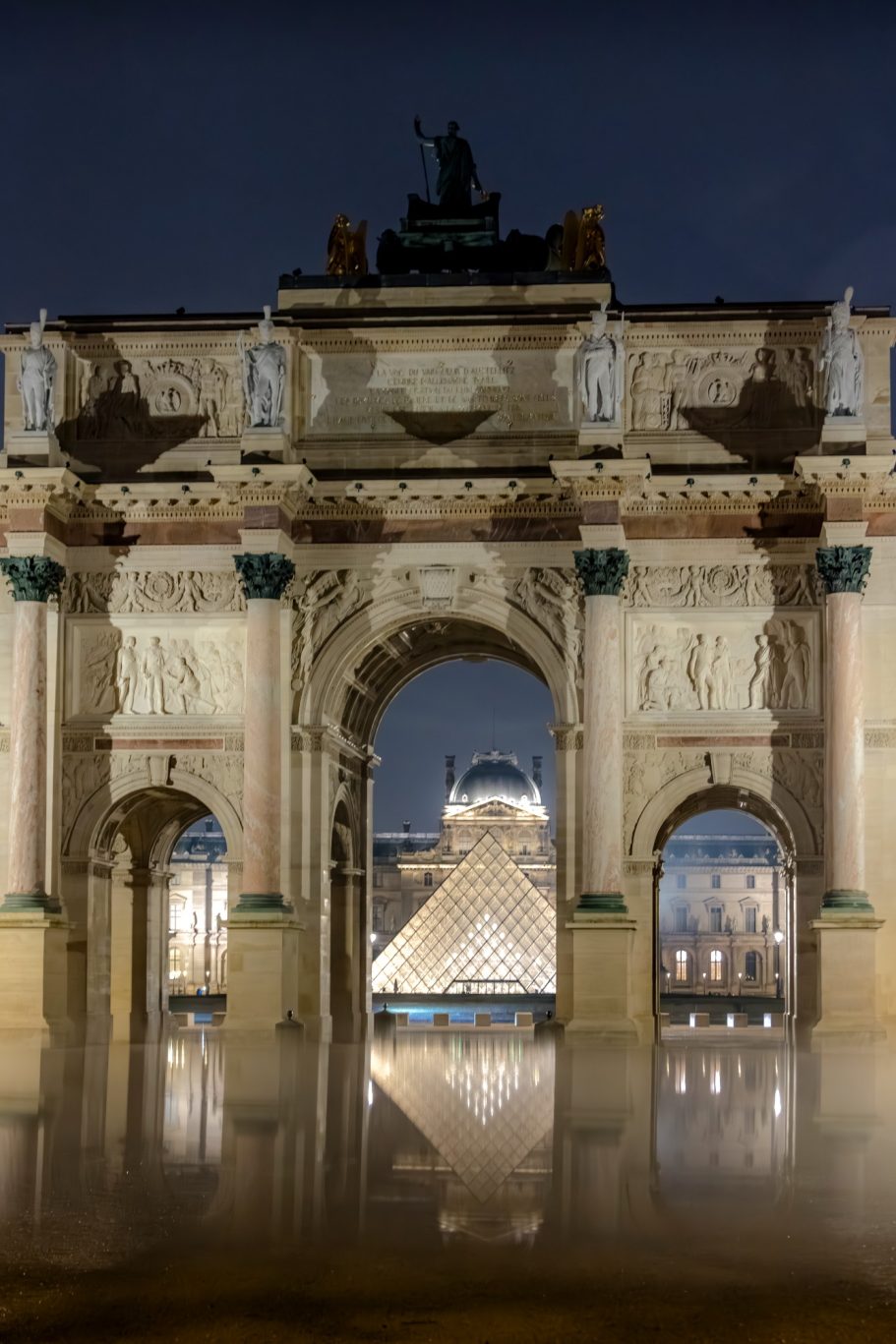 Arch with ornate decorations, illuminated, with the glass pyramid of the Louvre in the foreground.