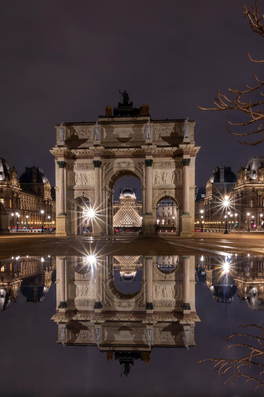 Arch with water reflection at night, surrounded by illuminated buildings.
